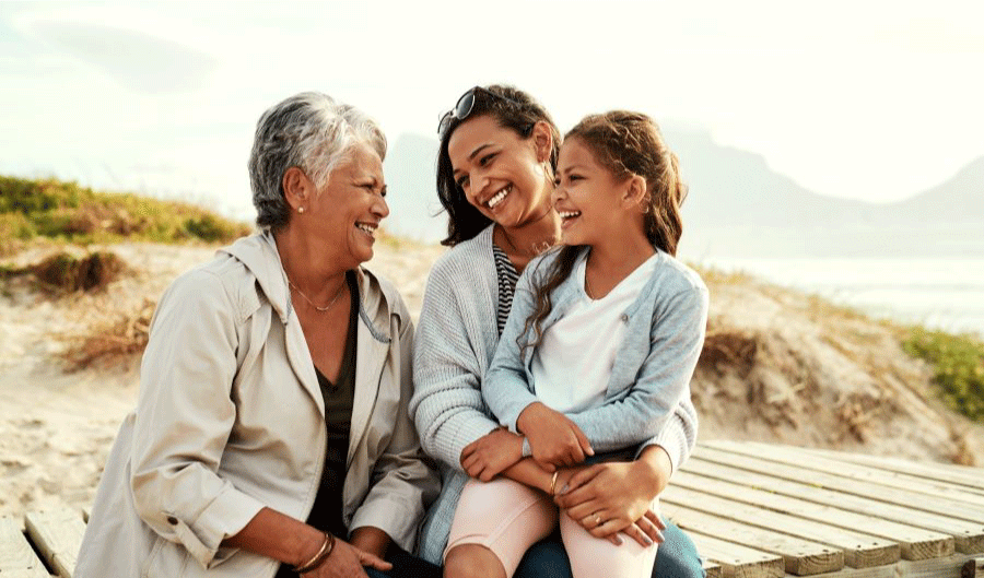 Grandmother, mother, and young daughter sit on beach, embracing and smiling at each other