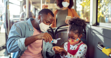 family of three sits on a city bus, dad puts hand sanitizer on daughter, all three wear masks