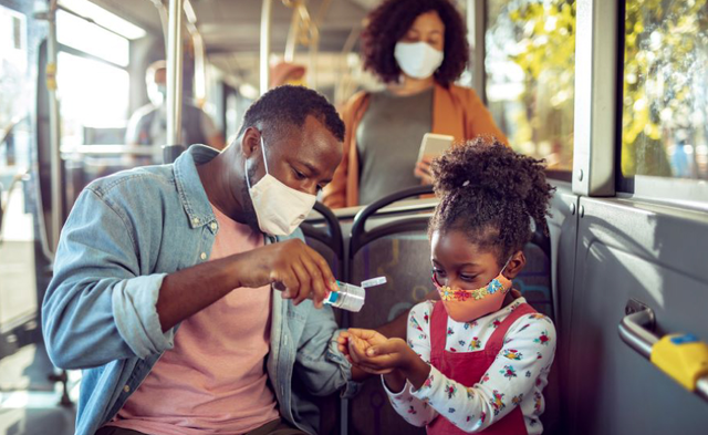family of three sits on a city bus, dad puts hand sanitizer on daughter, all three wear masks