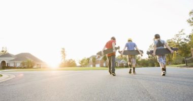 Three teenaged boys walking down the street toward the sunset, skateboards in hand