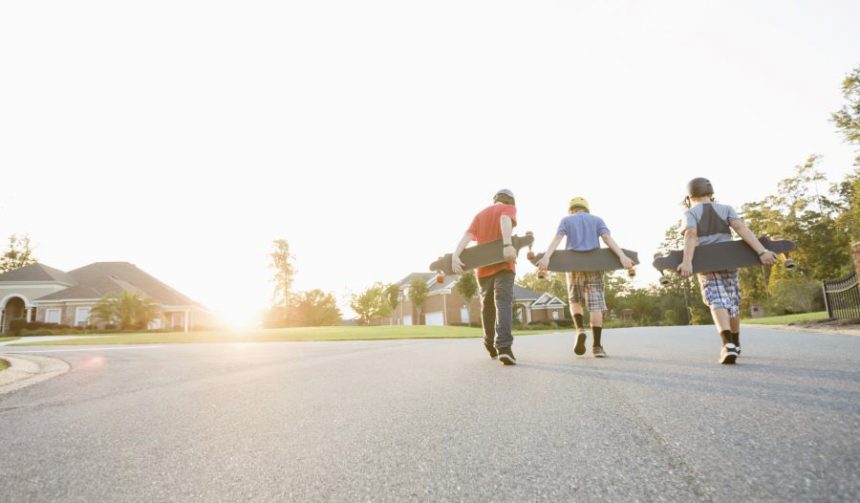 Three teenaged boys walking down the street toward the sunset, skateboards in hand