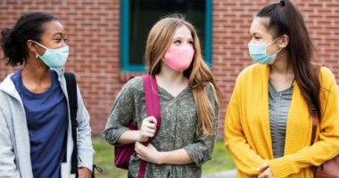 Three middle-school girls walk together outside of the school building, all three wearing backpacks and face masks