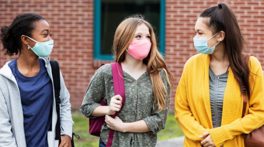 Three middle-school girls walk together outside of the school building, all three wearing backpacks and face masks
