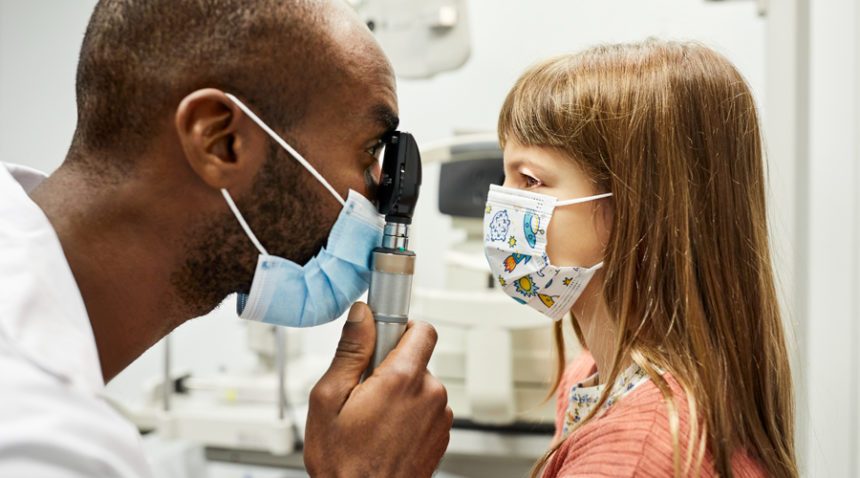 Provider checks eyes of little kid with flashlight, both wearing face masks
