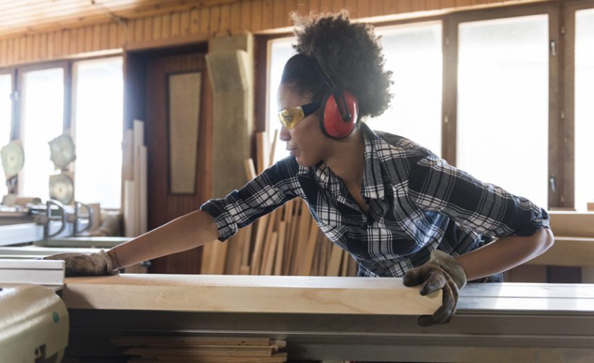 woman in a woodworking shop, working with equipment. She is wearing ear and eye protection.