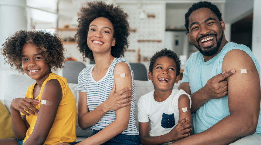 Family of four (Mom, Dad, two kids) sit on couch with sleeves rolled up, each smiling and pointing at their arms, where there is a bandage from when they each received a vaccine