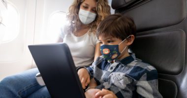 mother watches young son play with a tablet while sitting in airplane seats, both are wearing face masks