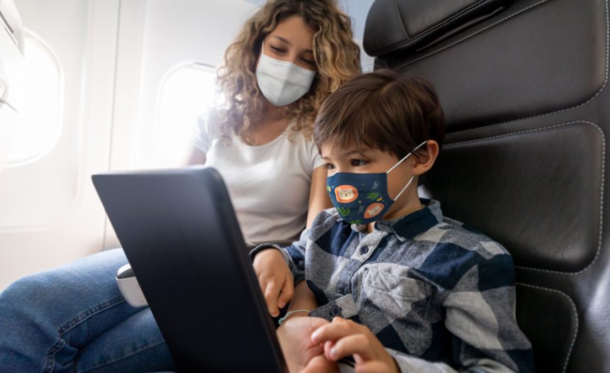 mother watches young son play with a tablet while sitting in airplane seats, both are wearing face masks