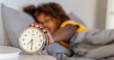 Out-of-focus child sleeping in bed puts hand on in-focus alarm clock
