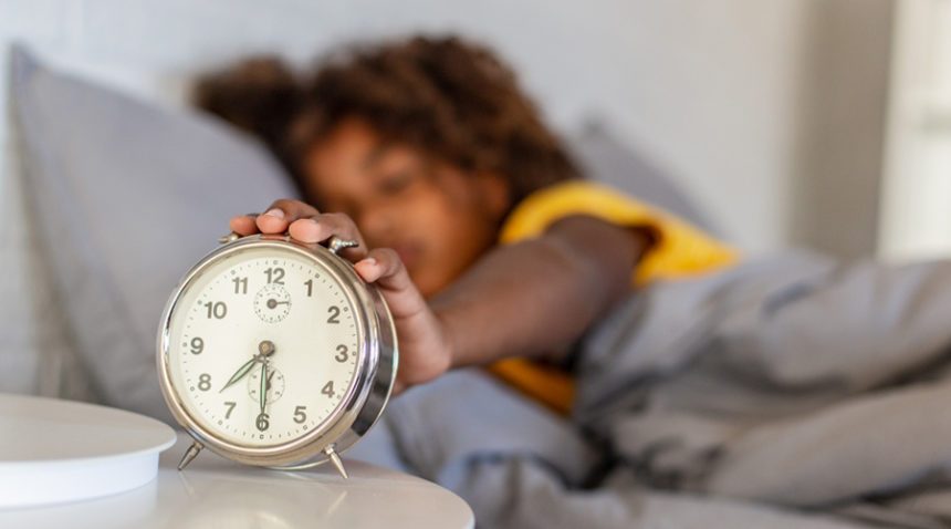 Out-of-focus child sleeping in bed puts hand on in-focus alarm clock