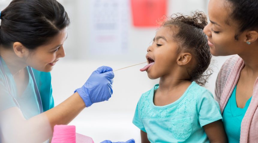 Little girl sticks out her tongue for a swab by a provider while her mom stands next to her.