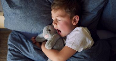 little boy yawns as he lies in bed, clutching a stuffed bunny