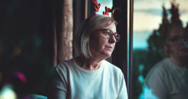 woman wearing a festive reindeer antler headband looks out of the window, a wistful, sad expression on her face