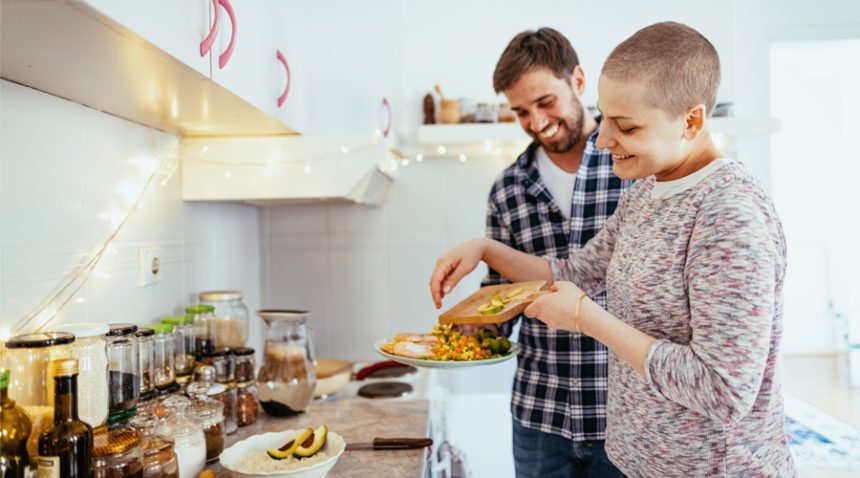male/female couple cooks dinner together in a kitchen. Female has a shaved head, indicating she is going through chemotherapy.