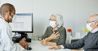 Older couple wearing face masks speaks to provider, also wearing a face mask, and seated at a desk