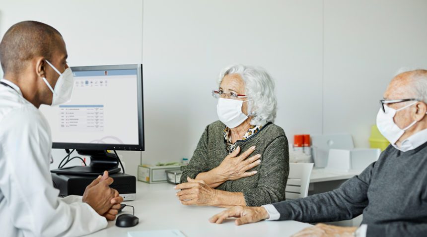Older couple wearing face masks speaks to provider, also wearing a face mask, and seated at a desk