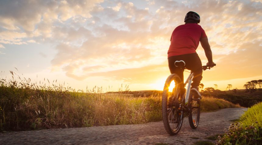 back of a man riding on a bicycle on a dirt path with grass surrounding, riding toward beautiful sunset