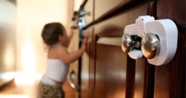close-up image of safety lock on cabinet handles, blurred in background is a busy toddler trying to open another cabinet door