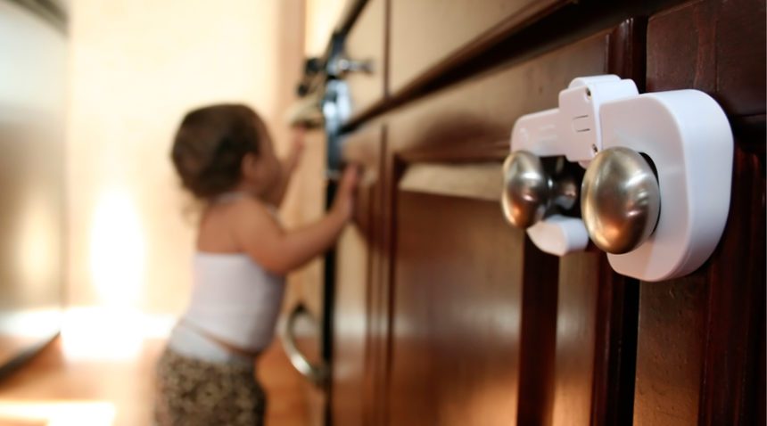 close-up image of safety lock on cabinet handles, blurred in background is a busy toddler trying to open another cabinet door