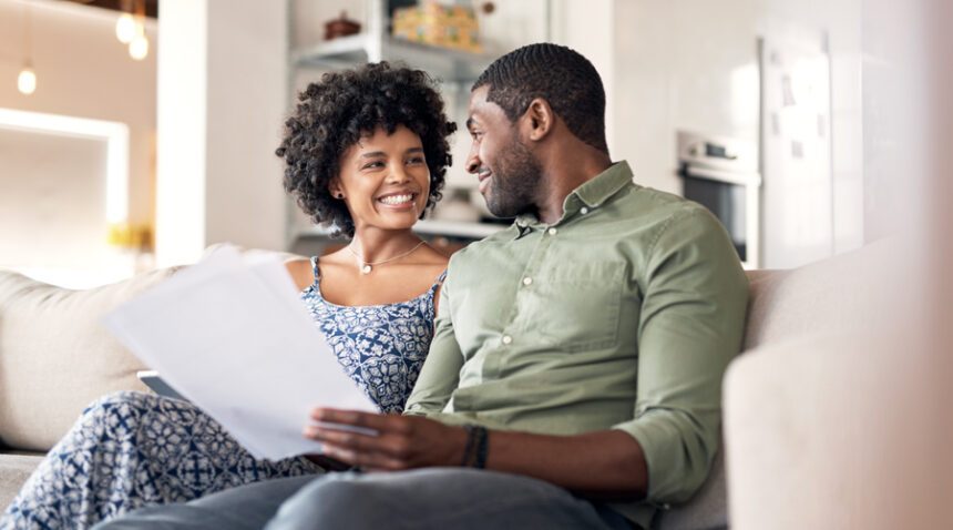 Young, happy couple sits on couch looking at each other, holding papers to show they are discussing paperwork