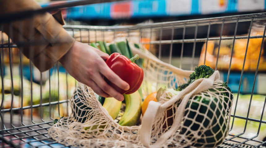 close-up image of a person putting a red bell pepper into a mesh bag filled with other vegetables in a grocery shopping cart