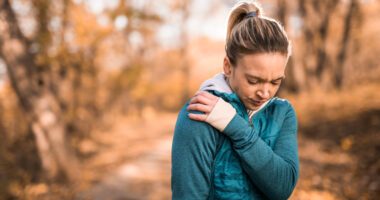 woman wearing winter exercise clothing, standing in the woods in fall or winter season, clutches shoulder in pain
