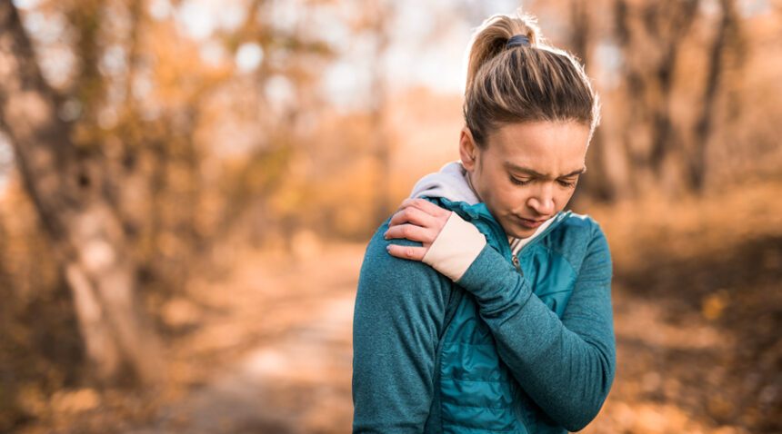 woman wearing winter exercise clothing, standing in the woods in fall or winter season, clutches shoulder in pain