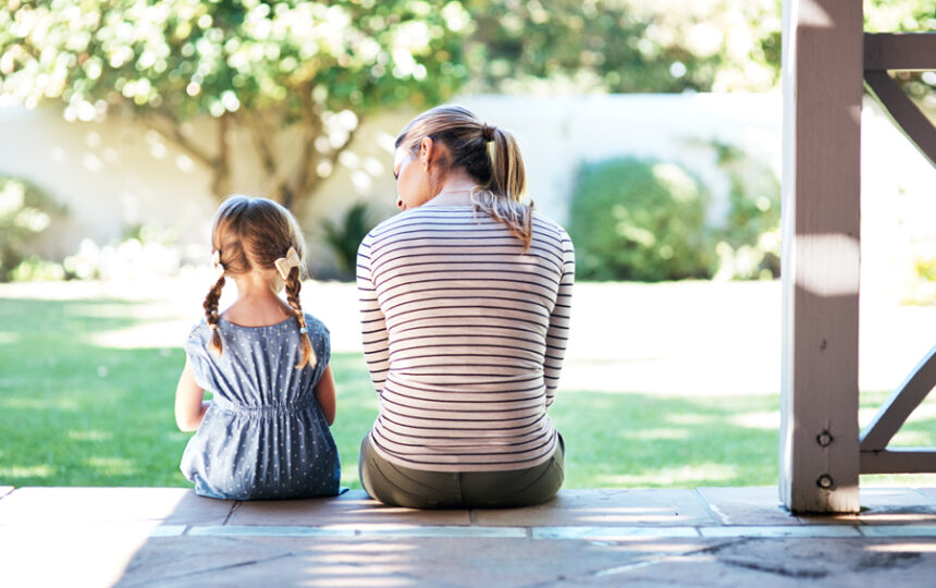 mom sits on porch with young daughter, their backs to the camera