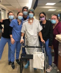 Lung transplant patient Sean Noble, in a hospital gown, mask, and using a walker, poses with this care team
