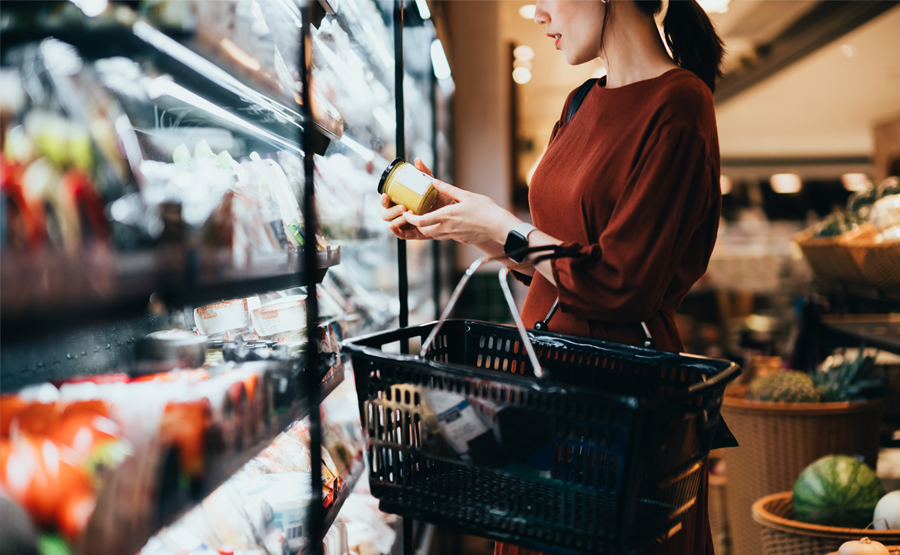 midshot of a woman looking at an item in the refrigerated section at the grocery store, grocery basket on arm.