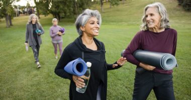 Two older women chat, carry yoga mats and water bottles as they walk in a park