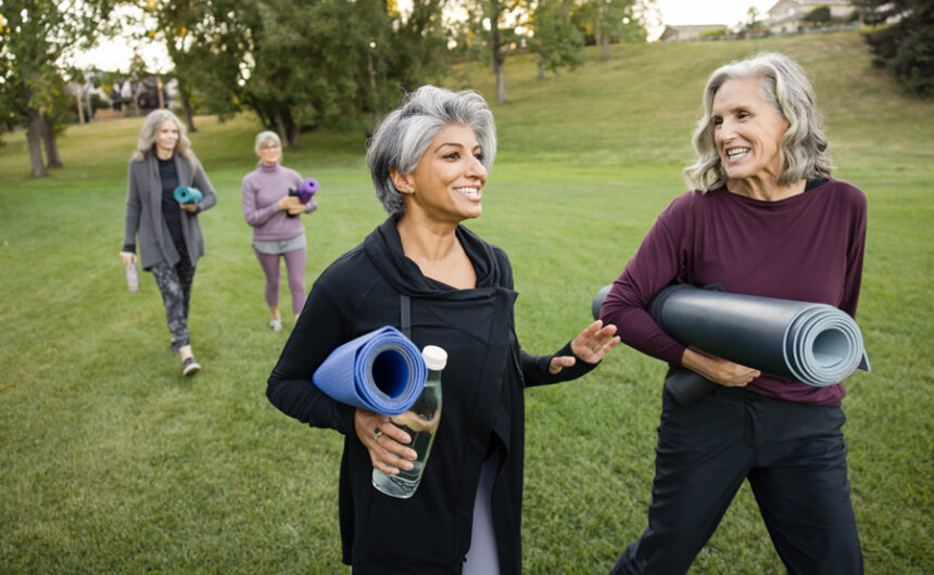 Two older women chat, carry yoga mats and water bottles as they walk in a park