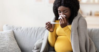 Pregnant woman sits on a couch, looks at a thermometer and wipes a tissue on her nose. She is covered in a blanket.