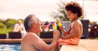 Grandfather and grandson smile while eating popsicles; grandson is sitting on edge of a pool, grandfather is in the pool.