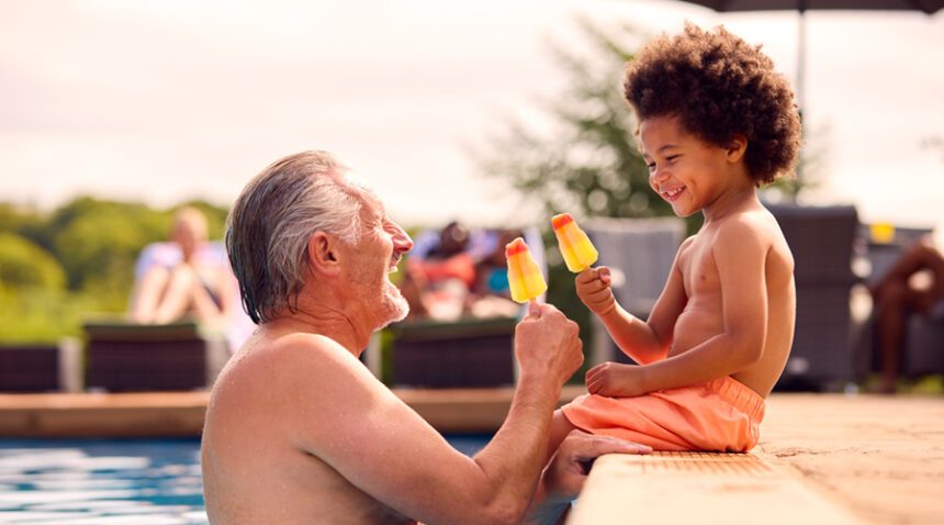 Grandfather and grandson smile while eating popsicles; grandson is sitting on edge of a pool, grandfather is in the pool.