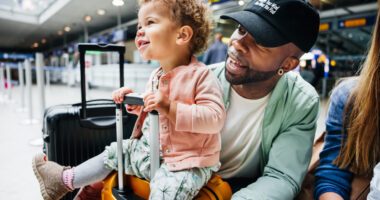 Father pushes toddler who is sitting on top of a rolling suitcase in an airport