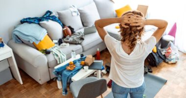 woman stands in a messy living room, hands on head in exasperation