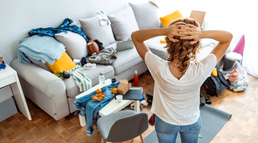 woman stands in a messy living room, hands on head in exasperation