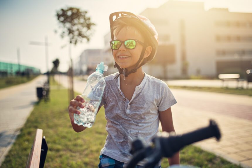 Boy sits on bicycle, wearing a helmet and sunglasses. He drinks a bottle of water. There is a sun flare in the top right of the photo, indicating a warm, sunny day.