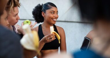 young woman in fitness apparel smiles while holding a banana she is eating