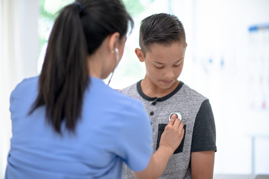 Provider, with back to camera, holds stethoscope up to young adolescent boy's chest to listen to his heart.