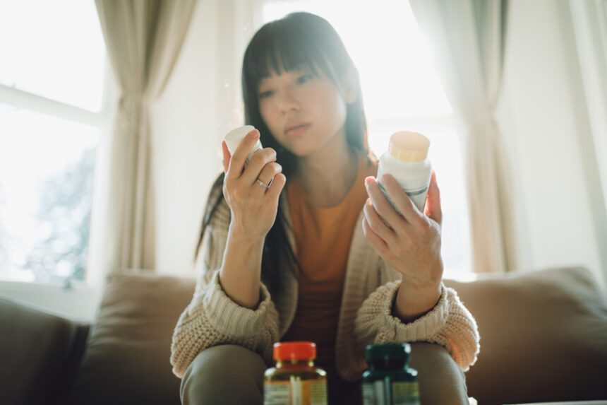 Young woman sits on couch, holding different medicine/vitamin bottles to read the labels