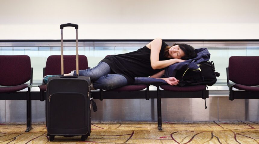 female traveler sleeps across a cluster of seats at an airport, uses her suitcase for a foot rest