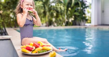 little kid wearing a swimsuit sits on a side of a pool, eating a slice of watermelon