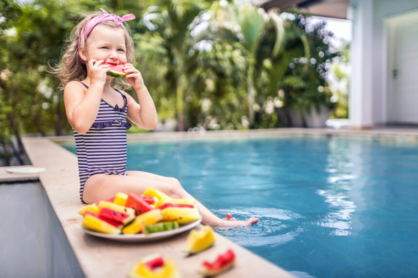 little kid wearing a swimsuit sits on a side of a pool, eating a slice of watermelon