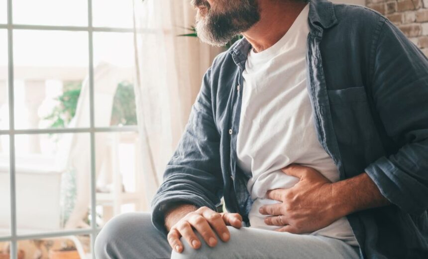 middle-aged man sits on couch, gripping his stomach in pain