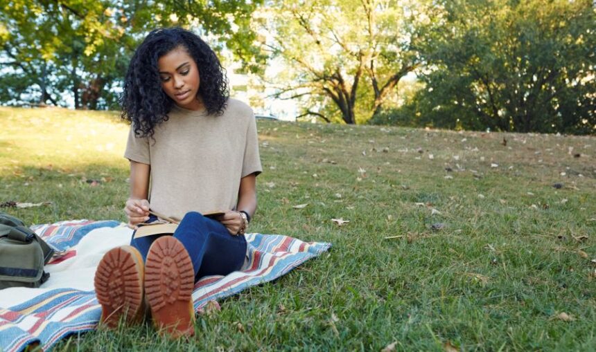 Woman sits outside in a nice shaded park, sits on blanket while reading a book