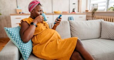 pregnant woman sits on couch, drinking team and looking at her smart phone