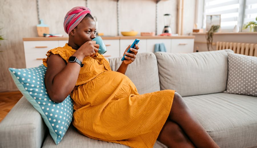 pregnant woman sits on couch, drinking team and looking at her smart phone