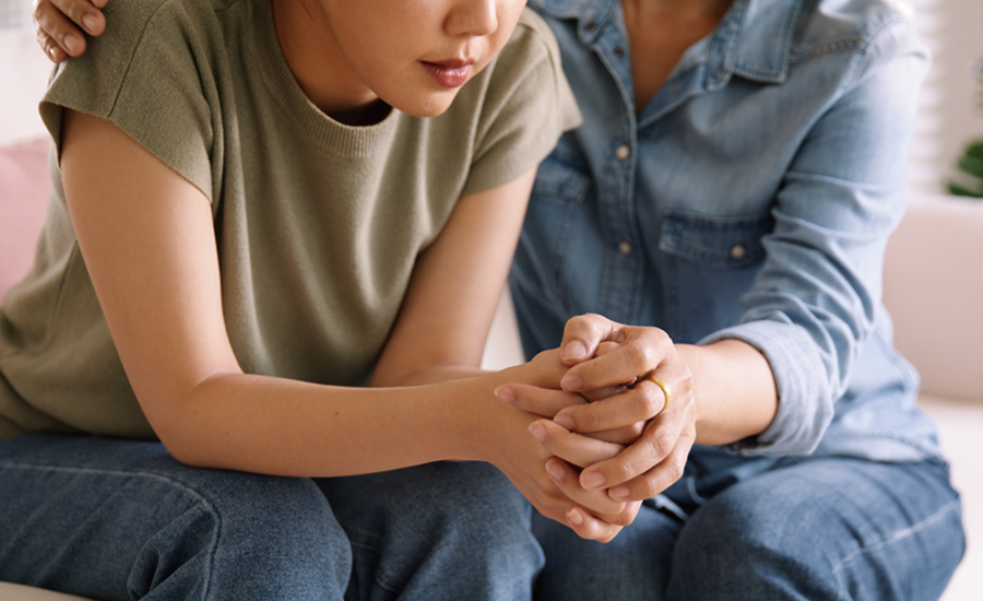 Mom sits with young adolescent on a couch, holding their hand and consoling them